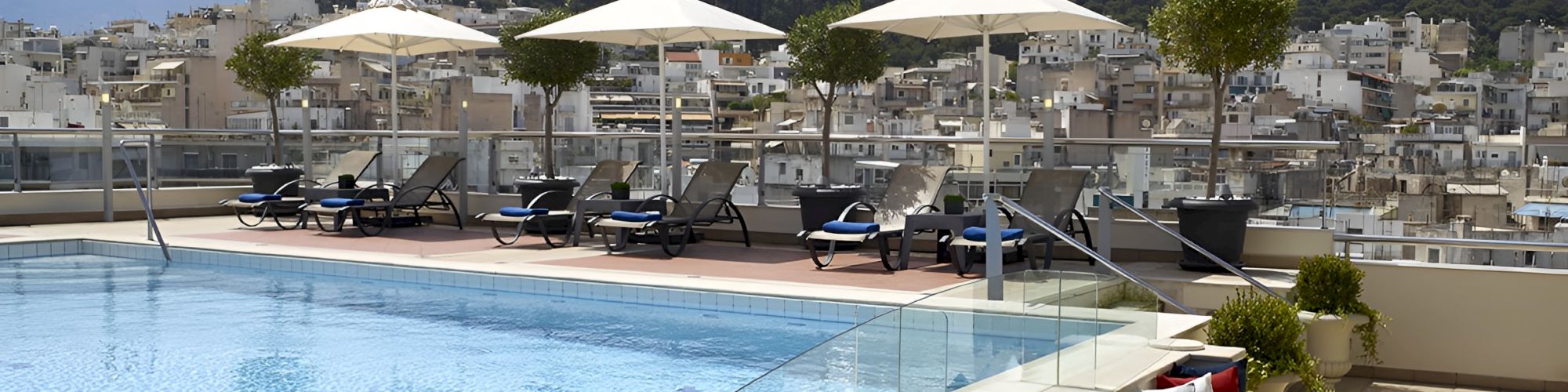 Rooftop swimming pool with lounge chairs and white umbrellas, overlooking a hillside town under a clear blue sky.