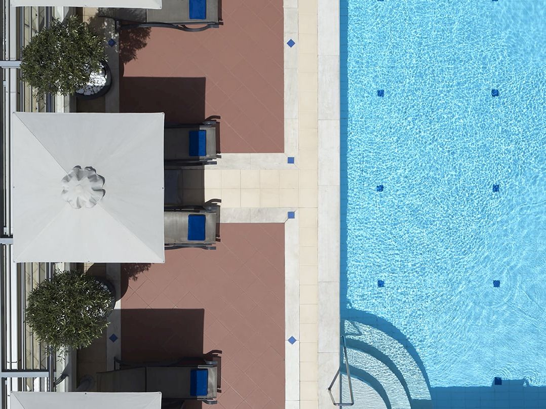 Blue swimming pool beside an outdoor patio with white umbrellas and lounge chairs in a sunlit hotel courtyard.