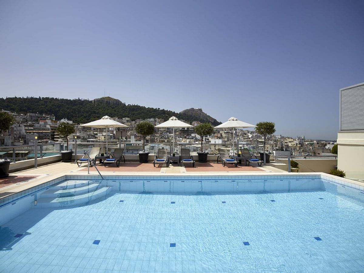 Rooftop swimming pool with lounge chairs and white umbrellas, overlooking a coastal town and green hills under a clear sky.