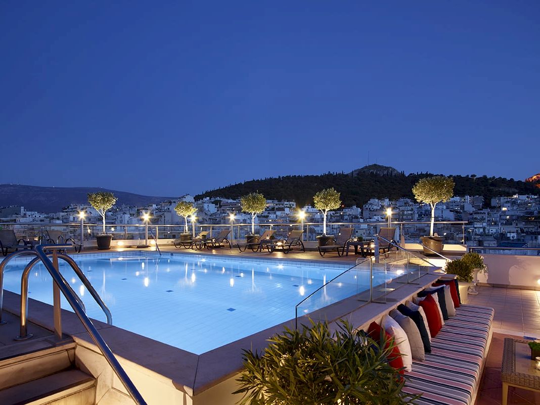 A rooftop hotel pool at dusk with lounge chairs, lights, and a hillside city view behind it.