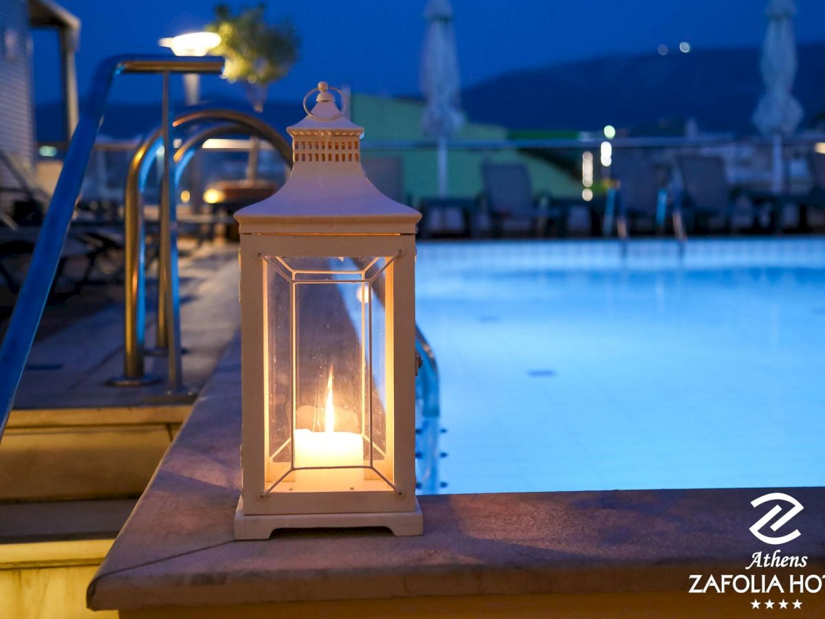 A glowing lantern sits on a poolside ledge at dusk, with a blue pool and blurred buildings in the background.