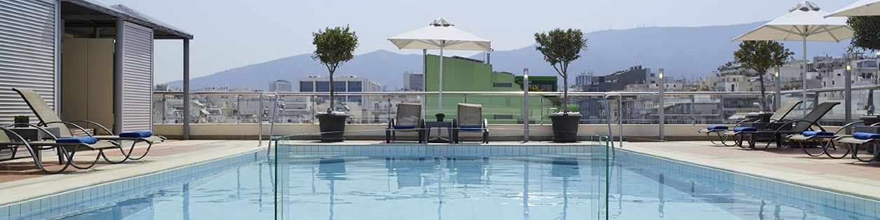 Rooftop pool with lounge chairs, umbrellas, and a cityscape view under a clear sky.