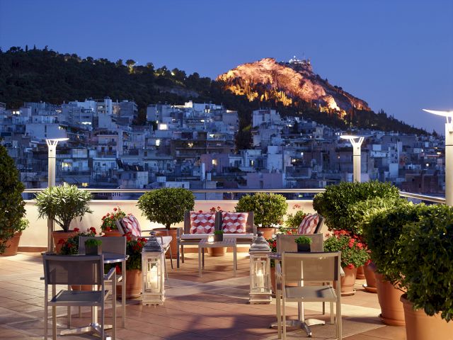 Rooftop terrace with potted greenery and lanterns overlooking a hillside city at dusk, with a torch-lit mountain behind.