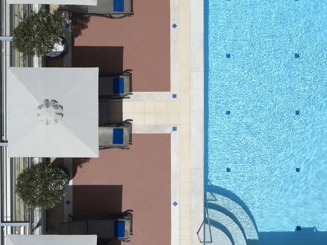 A bright outdoor pool with blue water beside lounge chairs, umbrellas, and potted plants in a tiled courtyard.