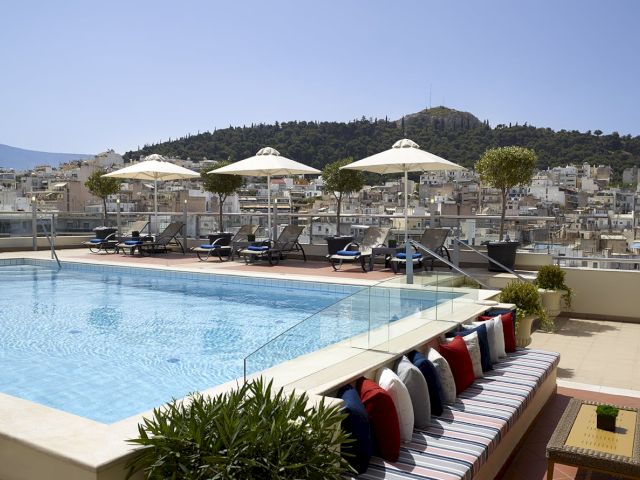 Rooftop hotel pool with sunbeds and white umbrellas, overlooking a hillside city under a clear blue sky.
