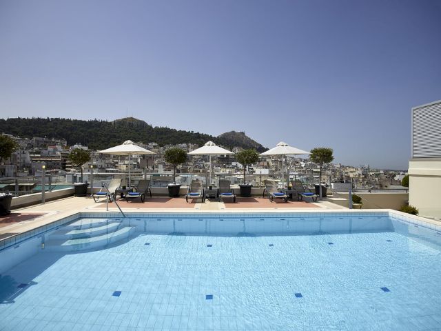 A rooftop hotel pool with sun loungers, white umbrellas, and a view of hillside city buildings under a clear blue sky.