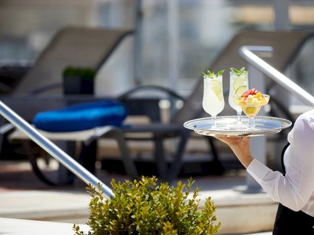 A server holds a silver tray with two colorful cocktails on an outdoor caf&eacute; patio, with chairs and tables blurred behind them.