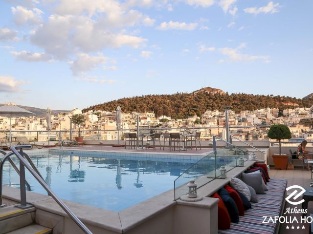 Rooftop hotel pool with loungers and umbrellas overlooks a hillside town under a bright blue sky with scattered clouds.