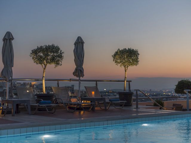 A rooftop pool at sunset with lounge chairs, blue umbrellas, small trees, and a city skyline in the distance.