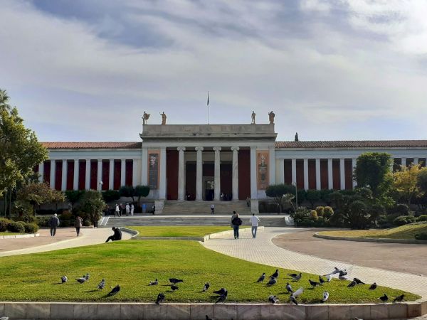A grand neoclassical building with tall columns, people on steps, landscaped garden and pigeons on the lawn under a cloudy sky.