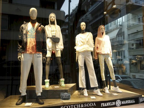 A clothing store window displays four mannequins in casual hoodies and jeans, with sneakers on a lit platform.