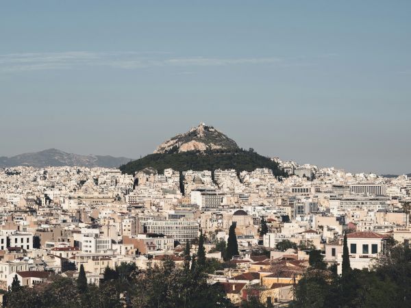 A sprawling city skyline surrounds a rocky hill crowned with a small fortress, beneath a clear blue sky.