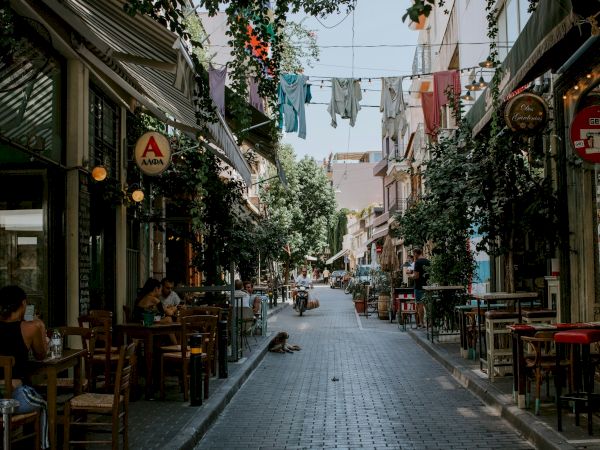 A leafy city alley with caf&eacute; tables, hanging string lights and clothes, and pedestrians strolling between storefronts, all bathed in daylight.
