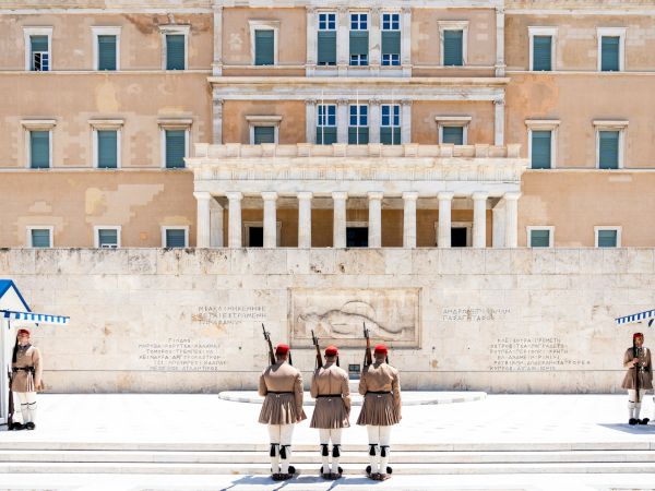 A grand neoclassical building fa&ccedil;ade with tall columns and windows, with uniformed soldiers standing guard in front of it.