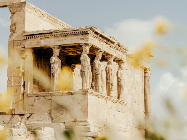 Ancient Greek temple ruins with carved statues and tall columns under a cloudy sky, with sunlight streaming through haze.