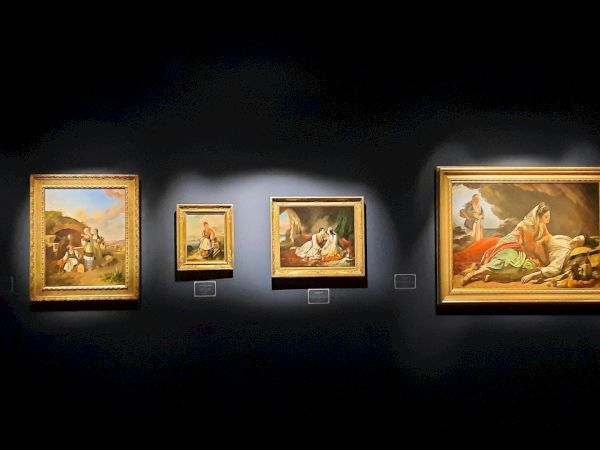 A dark gallery wall displays several framed paintings, with a person standing at the right viewing the artwork.