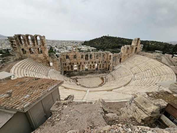 An ancient Roman amphitheater with curved stone seating, tall archways, and a view over a hillside city under cloudy skies.