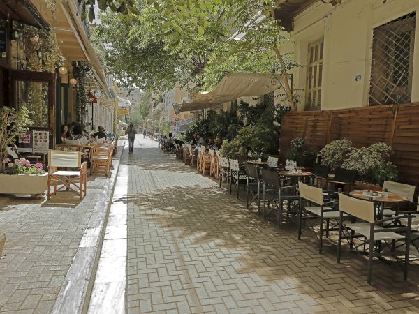 A sunny outdoor caf&eacute; street with tables and chairs along a narrow walkway, shaded by green trees and hanging awnings.