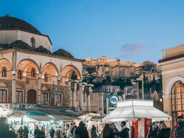 Ancient ruins and colonnaded buildings at a lively street market at dusk, with people walking beneath a blue sky.