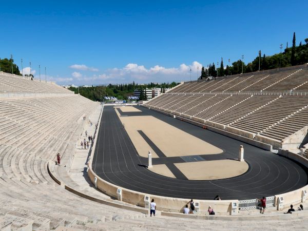 Ancient-style stone amphitheater with an empty track and tiered seats under a bright blue sky.