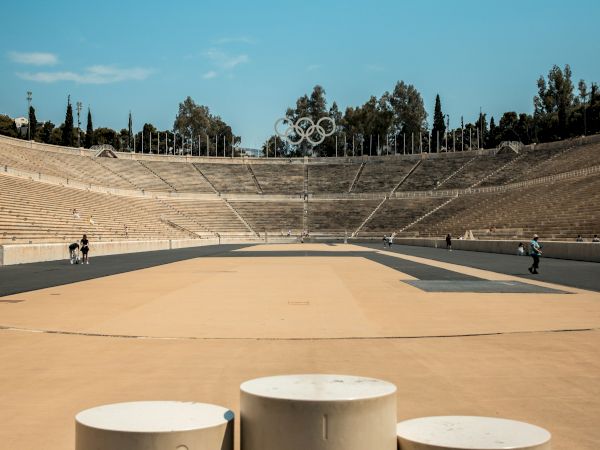 An Olympic stadium with empty stone bleachers and the Olympic rings centered on the field under a clear blue sky.