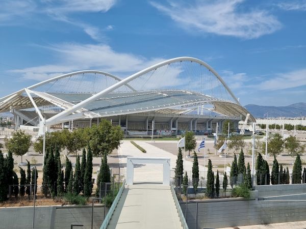 A modern stadium with sweeping white arches and a curved netted roof, seen under a blue sky with scattered clouds.