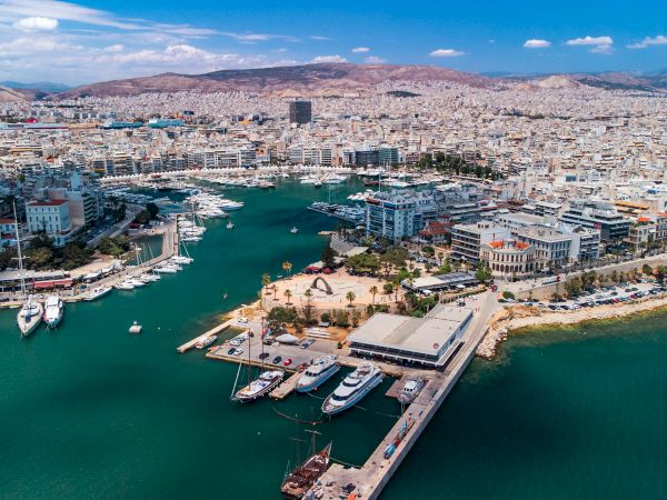 An aerial view of a busy harbor city with marinas and boats, surrounded by hills and mountains under a bright blue sky.