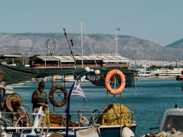 Boats are moored in a sunny harbor with an orange lifebuoy and flags, backed by hills and mountains.