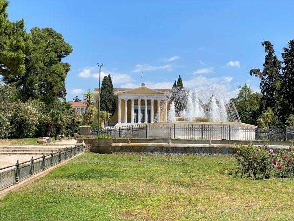 A sunny garden courtyard with a large fountain in front of a grand neoclassical building featuring the word Zappeion.
