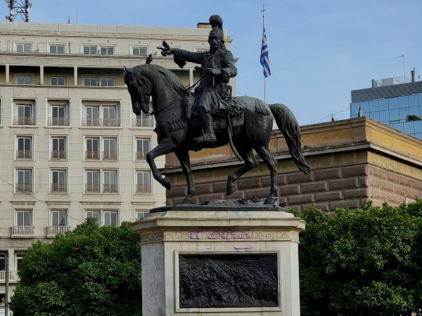 A bronze equestrian statue of a rider on horseback stands in a plaza beside a Greek flag and city buildings.