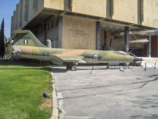 A camouflaged military jet sits outside a hangar, with serial markings visible on its fuselage.