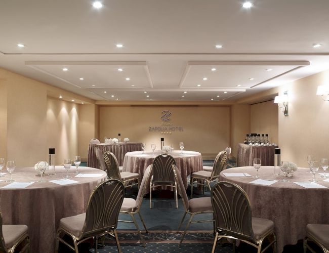 A hotel banquet meeting room with round tables, chairs, and a &ldquo;Zafolia Hotel&rdquo; sign under ceiling spotlights.