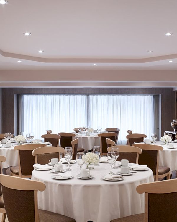A formal banquet dining room with tables set in white linens, glassware, and chairs, lit by recessed ceiling lights.