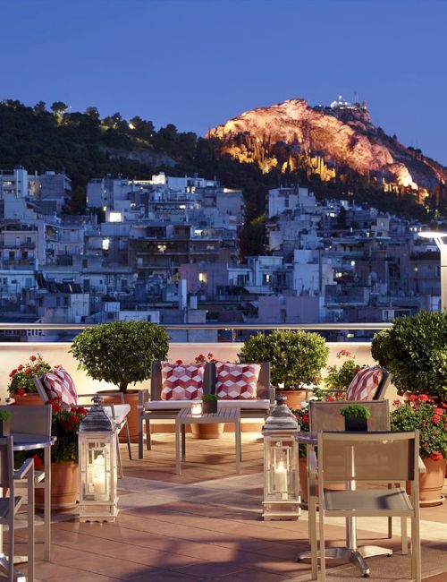A rooftop terrace with potted plants and lanterns overlooks a glowing hillside town at dusk.
