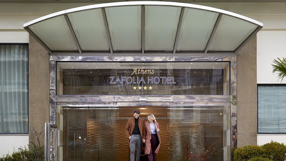 Entrance of Athens Zafolia Hotel with glass doors and two guests walking inside.