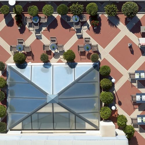 An outdoor rooftop patio with neatly spaced round trees, caf&eacute; tables, and a large glass structure in the center.
