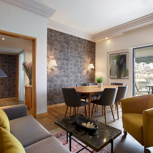A modern living-dining room with sofa, coffee table, wooden dining set, lamps, and large windows overlooking a town.