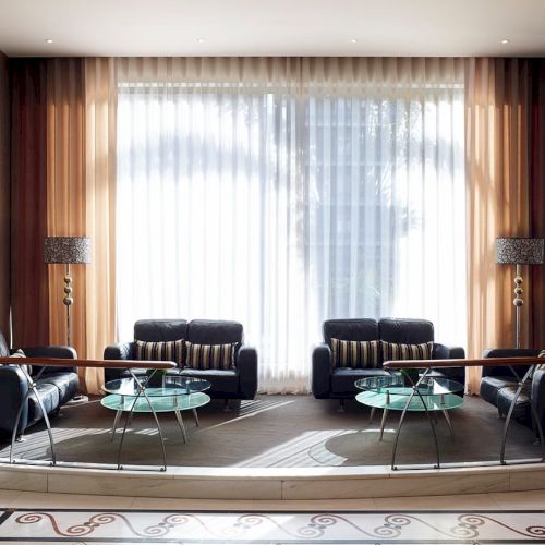 A modern living room with two dark sofas, glass round coffee tables, floor lamps, and sheer curtains framing a large window.