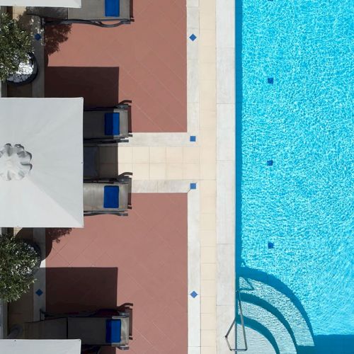 Sun loungers and a bright blue swimming pool beside hotel buildings in a quiet courtyard walkway.