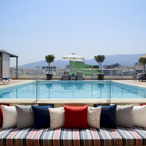 Rooftop hotel pool with sun loungers, umbrellas, and cushioned seating overlooking a city and mountains in clear daylight.