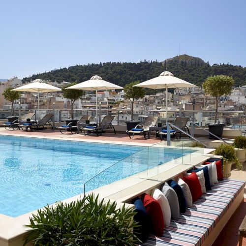 A rooftop hotel pool with sun loungers and white umbrellas, overlooking a hillside town under a clear blue sky.