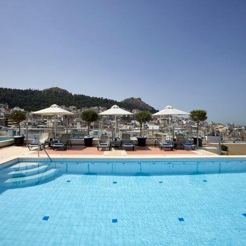 Rooftop hotel pool with blue water, loungers under white umbrellas, and a hillside city skyline in the background.