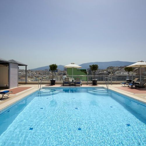 A bright rooftop swimming pool with lounge chairs and umbrellas, overlooking a city and distant hills.