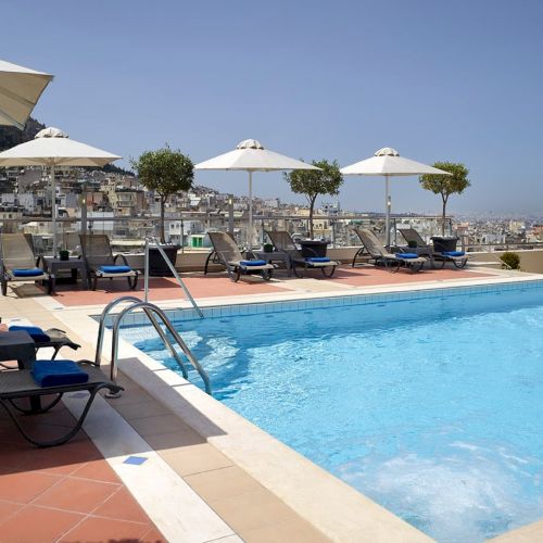 Rooftop hotel pool with blue water, lounge chairs, and white umbrellas overlooking a city skyline under a clear blue sky.
