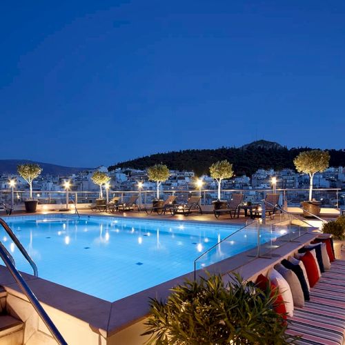 A rooftop swimming pool at dusk with lounge chairs, lit lamps, and a hillside town under a deep blue sky.