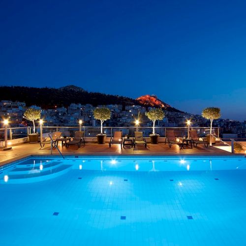 An outdoor rooftop swimming pool at dusk with lounge chairs, lit planters, and a hillside town under a deep blue sky.