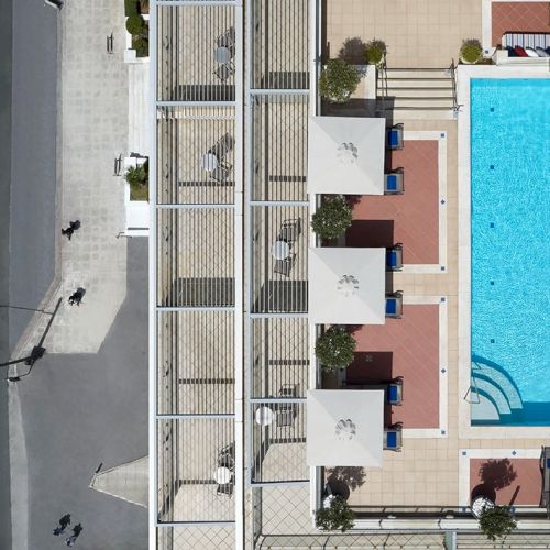 Aerial view of a building courtyard with a large bright blue swimming pool, sun loungers, and a street beside it.