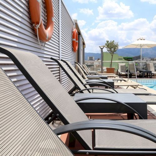 A rooftop hotel pool with blue water, sun loungers, umbrellas, and city views under a bright sky.