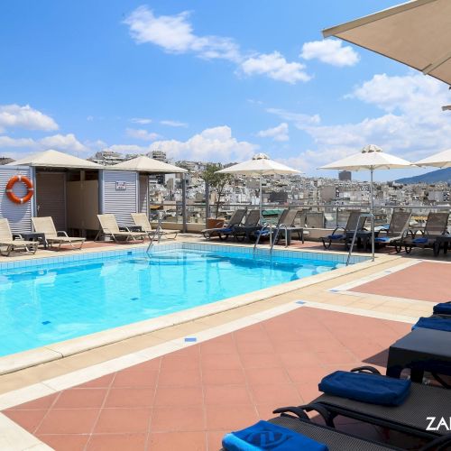 Rooftop hotel pool with sun loungers, umbrellas, and a view over the city under a blue sky.