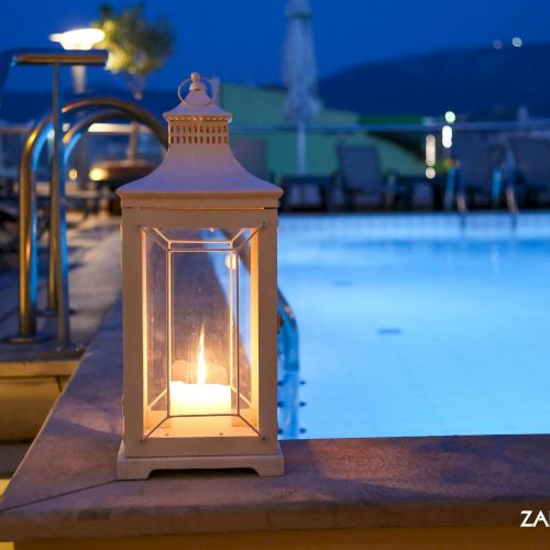 A glowing lantern on a poolside terrace at dusk, with the blue hotel pool and buildings softly blurred in the background.